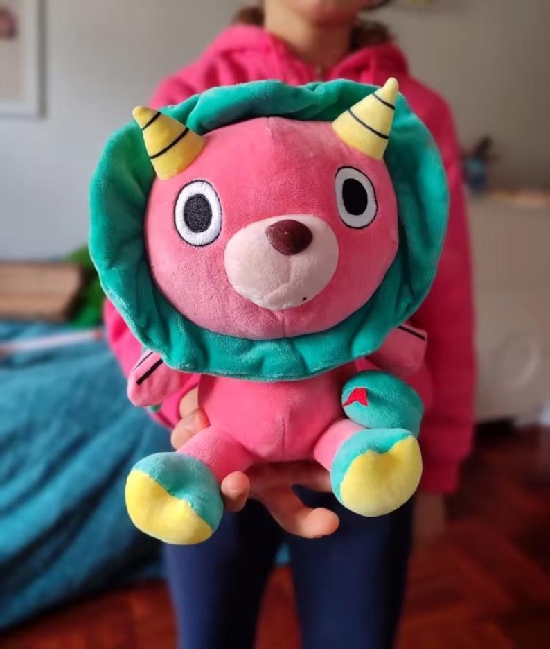Op de afbeelding: Een roze pluche speelgoed met een groene kap en gele hoorns. Het speelgoed heeft grote zwarte ogen en een bruine neus.