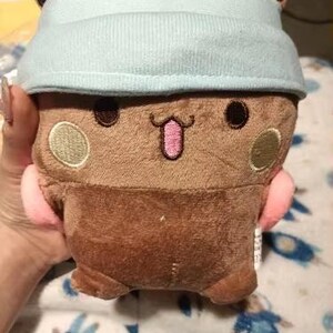 Puede incluir: Un juguete de peluche marr&oacute;n con un gorro azul claro. El juguete tiene una cara sonriente con mejillas rosadas y una lengua rosa que sobresale. El juguete tiene una nariz marr&oacute;n claro y dos ojos marrones.