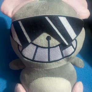 Puede incluir: Un peluche de rat&oacute;n gris con orejas y pies rosas, con gafas de sol negras. El rat&oacute;n tiene una gran sonrisa cosida y una barriga blanca. El juguete est&aacute; dise&ntilde;ado para parecer que lleva gafas de sol.