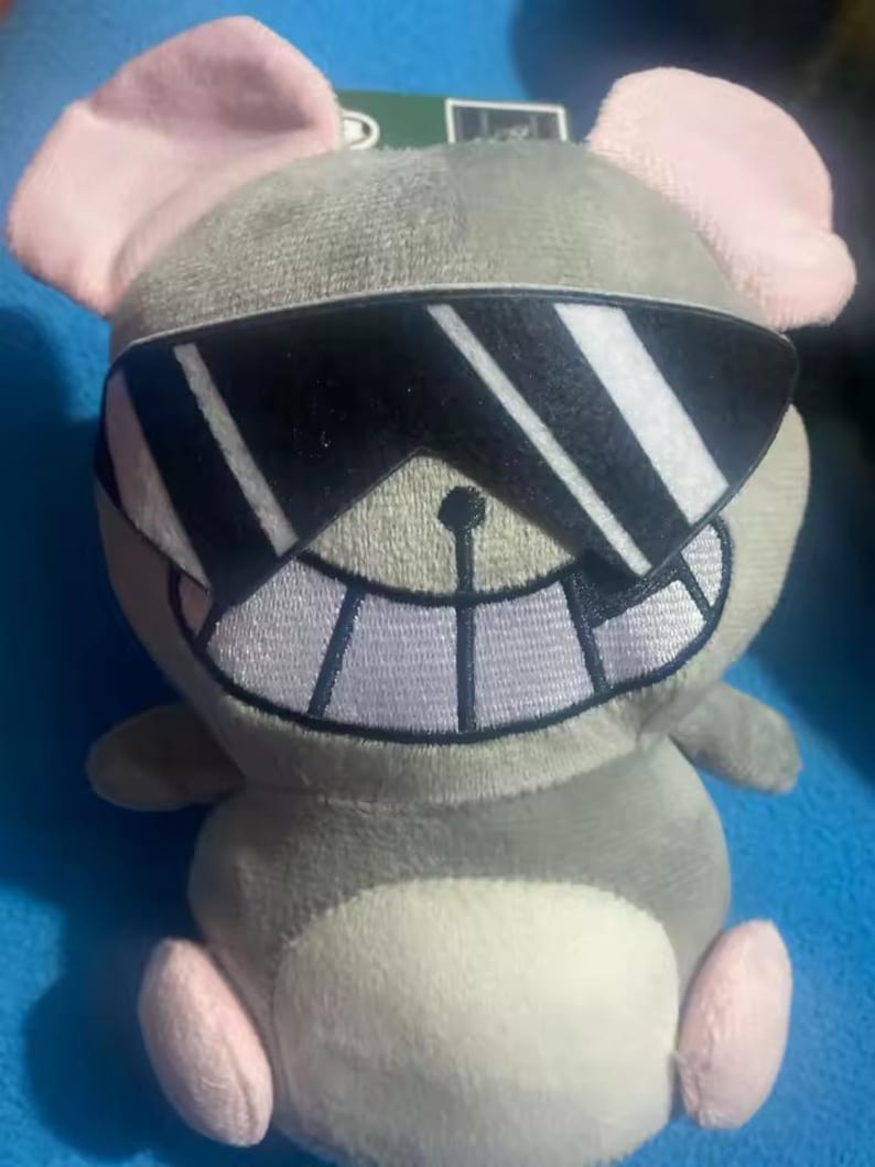 Puede incluir: Un peluche de rat&oacute;n gris con orejas y pies rosas, con gafas de sol negras. El rat&oacute;n tiene una gran sonrisa cosida y una barriga blanca. El juguete est&aacute; dise&ntilde;ado para parecer que lleva gafas de sol.
