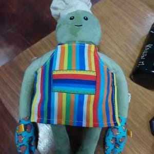 Puede incluir: Un peluche verde con un gorro de chef blanco, un delantal a rayas de colores y manoplas azules con un estampado marr&oacute;n y amarillo. El juguete tiene una cara sonriente y est&aacute; dise&ntilde;ado para parecer un chef.