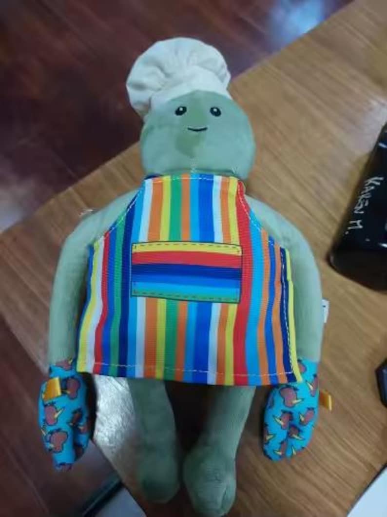 Puede incluir: Un peluche verde con un gorro de chef blanco, un delantal a rayas de colores y manoplas azules con un estampado marr&oacute;n y amarillo. El juguete tiene una cara sonriente y est&aacute; dise&ntilde;ado para parecer un chef.