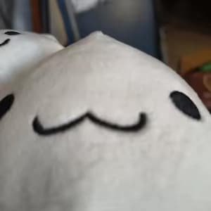 Puede incluir: Un juguete de peluche blanco con ojos bordados negros y una boca curvada. El juguete tiene una forma redondeada y una peque&ntilde;a parte superior puntiaguda. El fondo est&aacute; borroso, lo que sugiere un entorno interior.