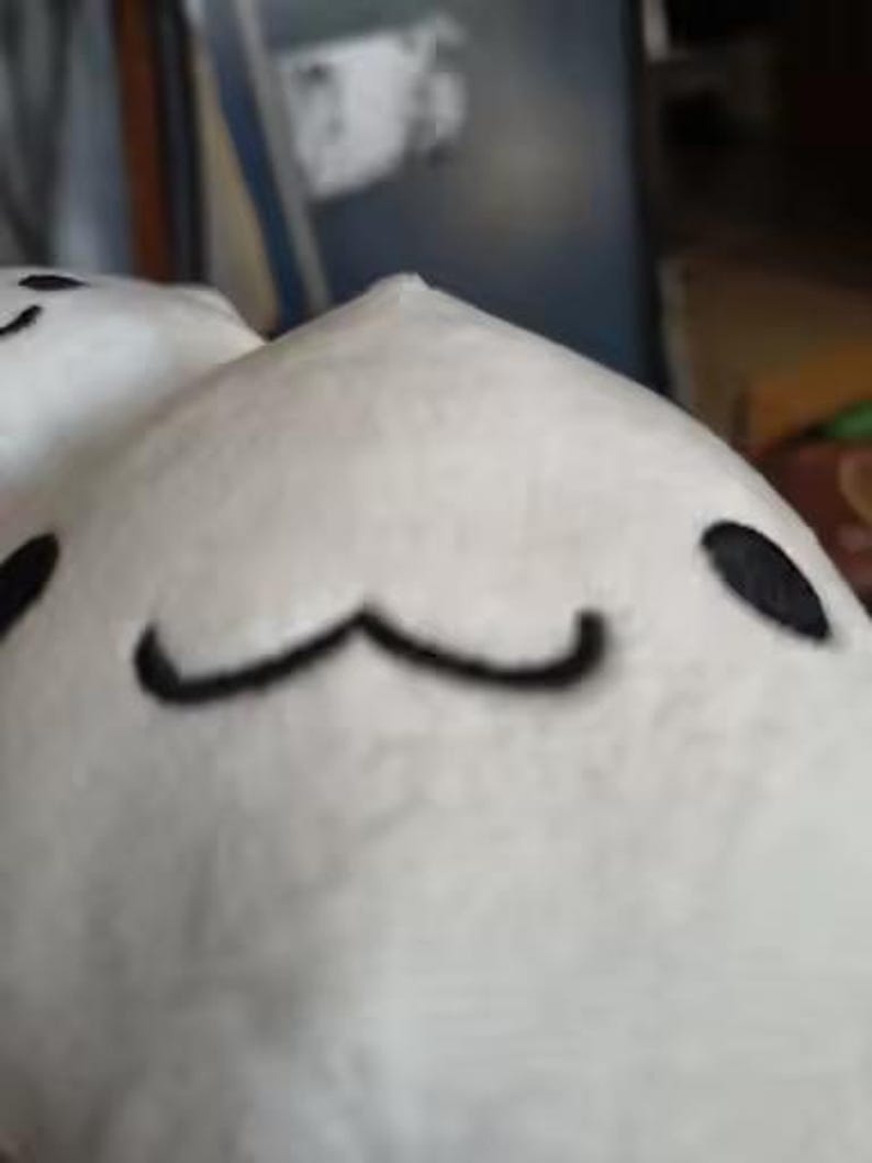 Puede incluir: Un juguete de peluche blanco con ojos bordados negros y una boca curvada. El juguete tiene una forma redondeada y una peque&ntilde;a parte superior puntiaguda. El fondo est&aacute; borroso, lo que sugiere un entorno interior.