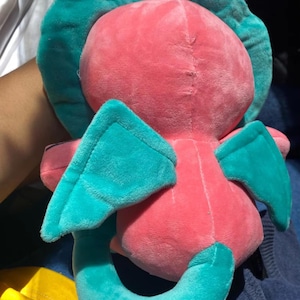 Op de afbeelding: Een pluche speelgoed in de vorm van een roze draak met turquoise vleugels en staart. Het speelgoed heeft een klein wit touwtje aan de bovenkant van zijn hoofd.