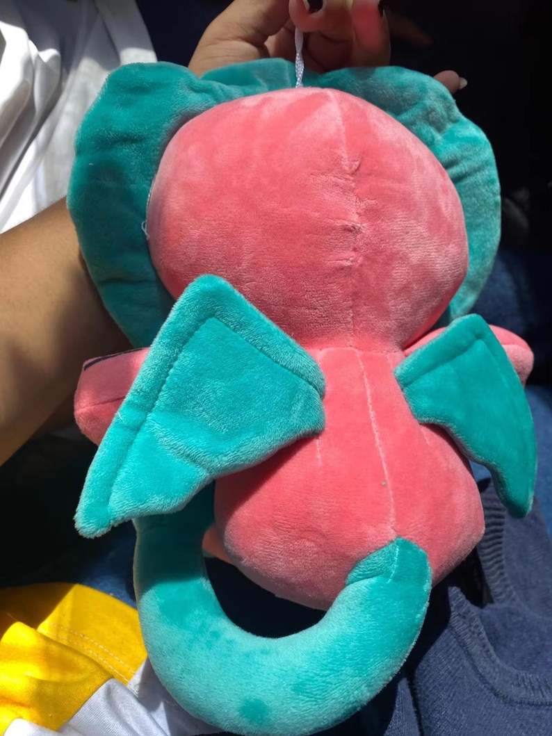 Op de afbeelding: Een pluche speelgoed in de vorm van een roze draak met turquoise vleugels en staart. Het speelgoed heeft een klein wit touwtje aan de bovenkant van zijn hoofd.