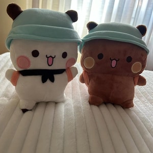 Puede incluir: Dos peluches con orejas marrones y sombreros azules. El peluche de la izquierda es blanco con mejillas rosadas y una corbata de lazo negra. El peluche de la derecha es marr&oacute;n con mejillas amarillas y una corbata de lazo marr&oacute;n.