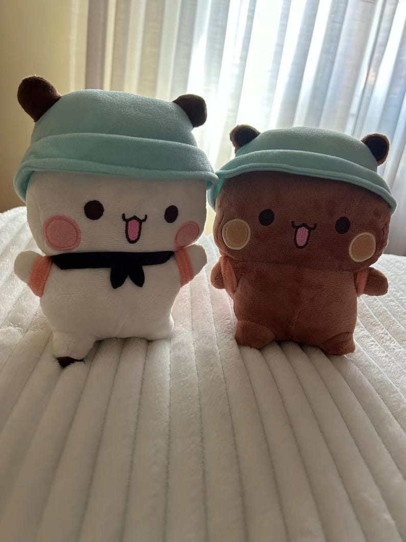 Puede incluir: Dos peluches con orejas marrones y sombreros azules. El peluche de la izquierda es blanco con mejillas rosadas y una corbata de lazo negra. El peluche de la derecha es marr&oacute;n con mejillas amarillas y una corbata de lazo marr&oacute;n.