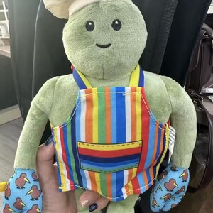 Puede incluir: Un personaje de peluche verde con un gorro de chef y un delantal a rayas de colores. El personaje tiene guantes azules con un estampado de kiwi. Un juguete suave y tierno para ni&ntilde;os.