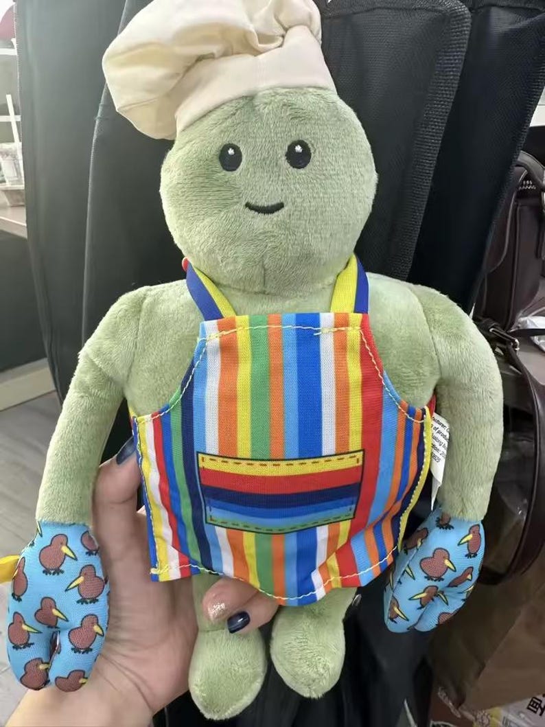 Puede incluir: Un personaje de peluche verde con un gorro de chef y un delantal a rayas de colores. El personaje tiene guantes azules con un estampado de kiwi. Un juguete suave y tierno para ni&ntilde;os.