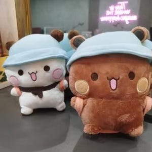 Puede incluir: Dos peluches, uno blanco con un sombrero azul y el otro marr&oacute;n con un sombrero azul. Ambos peluches tienen caras de dibujos animados lindas con ojos grandes y sonrisas.