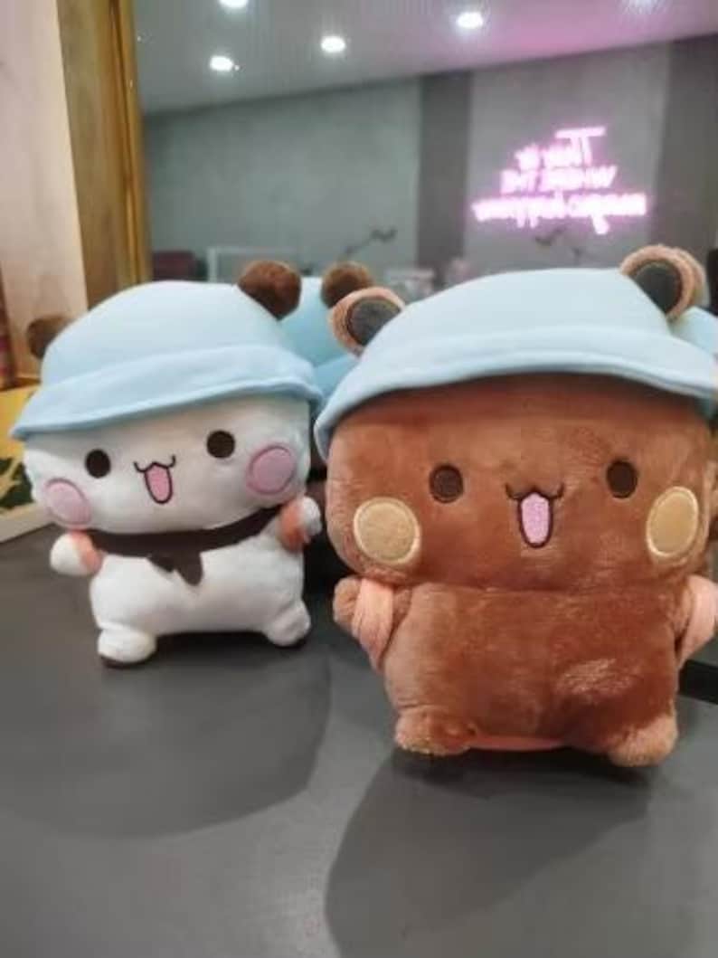 Puede incluir: Dos peluches, uno blanco con un sombrero azul y el otro marr&oacute;n con un sombrero azul. Ambos peluches tienen caras de dibujos animados lindas con ojos grandes y sonrisas.
