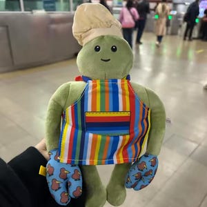 Puede incluir: Una tortuga de peluche verde con un gorro de chef, un delantal a rayas de colores y manoplas azules con diseños de aves kiwi. La tortuga tiene una cara sonriente. Un juguete suave y divertido.