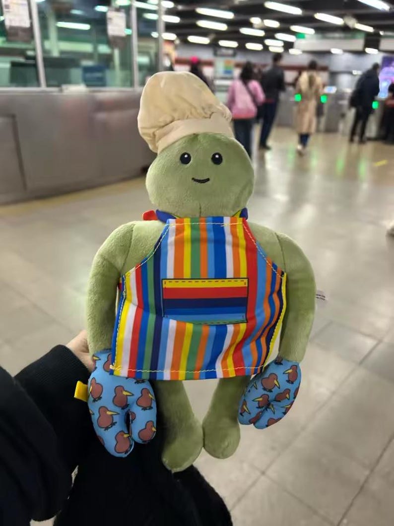Puede incluir: Una tortuga de peluche verde con un gorro de chef, un delantal a rayas de colores y manoplas azules con dise&ntilde;os de aves kiwi. La tortuga tiene una cara sonriente. Un juguete suave y divertido.