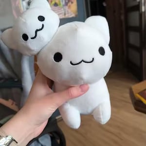 Puede incluir: Un peluche blanco con forma de gato con una cara sonriente y ojos negros. El juguete tiene una cabeza de gato m&aacute;s peque&ntilde;a adjunta en la parte superior. El peluche es sostenido por una persona.