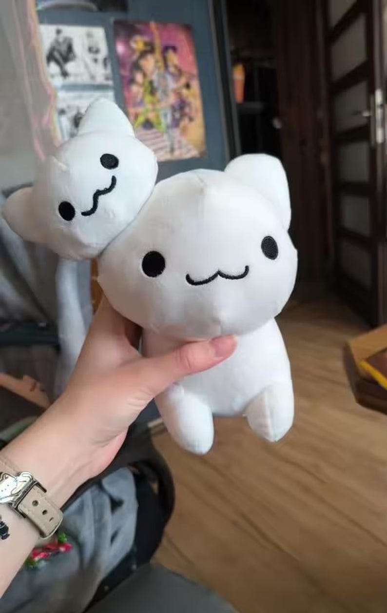 Puede incluir: Un peluche blanco con forma de gato con una cara sonriente y ojos negros. El juguete tiene una cabeza de gato m&aacute;s peque&ntilde;a adjunta en la parte superior. El peluche es sostenido por una persona.