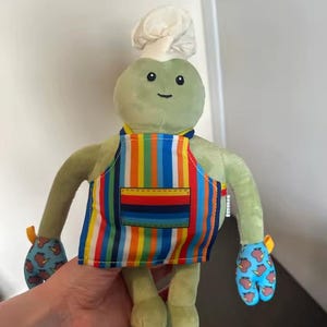 Puede incluir: Un peluche de rana verde con gorro de chef y delantal a rayas. La rana tiene una cara sonriente y manoplas con fondo azul y dise&ntilde;o de manzanas rojas.