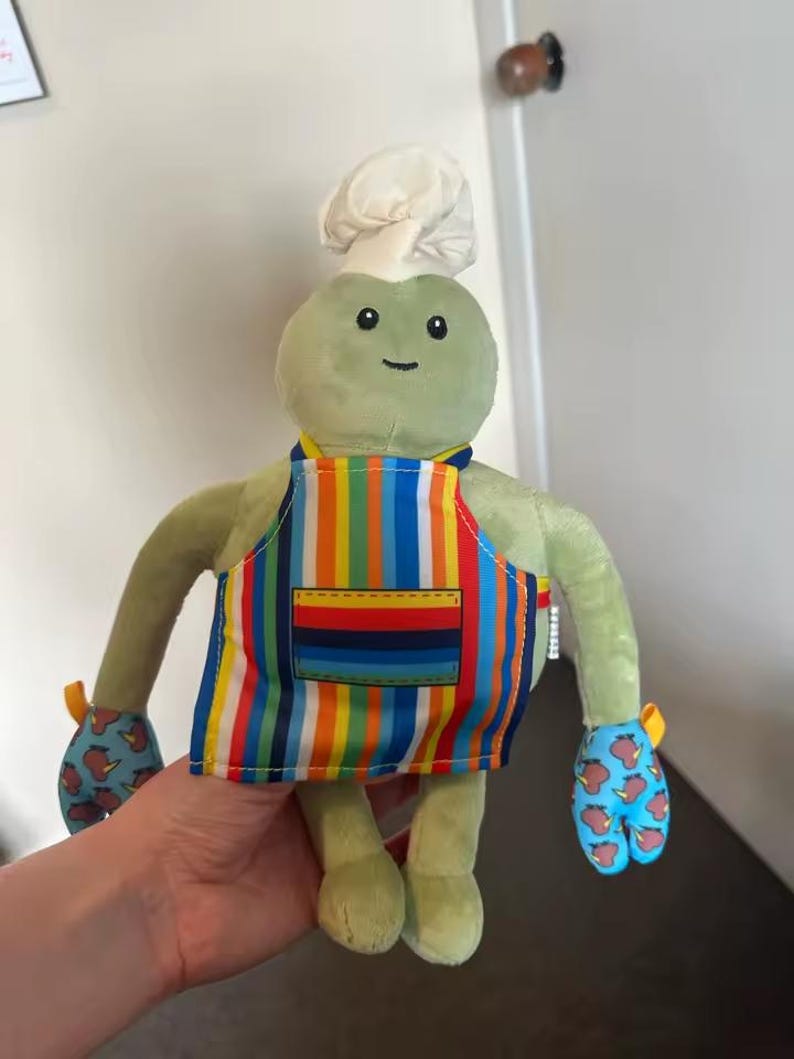 Puede incluir: Un peluche de rana verde con gorro de chef y delantal a rayas. La rana tiene una cara sonriente y manoplas con fondo azul y dise&ntilde;o de manzanas rojas.