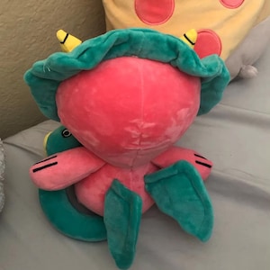Op de afbeelding: Een roze en groen pluche speelgoed met een groot hoofd en een klein lichaam. Het speelgoed heeft een groene hoed met gele accenten en een lange, gekrulde staart.