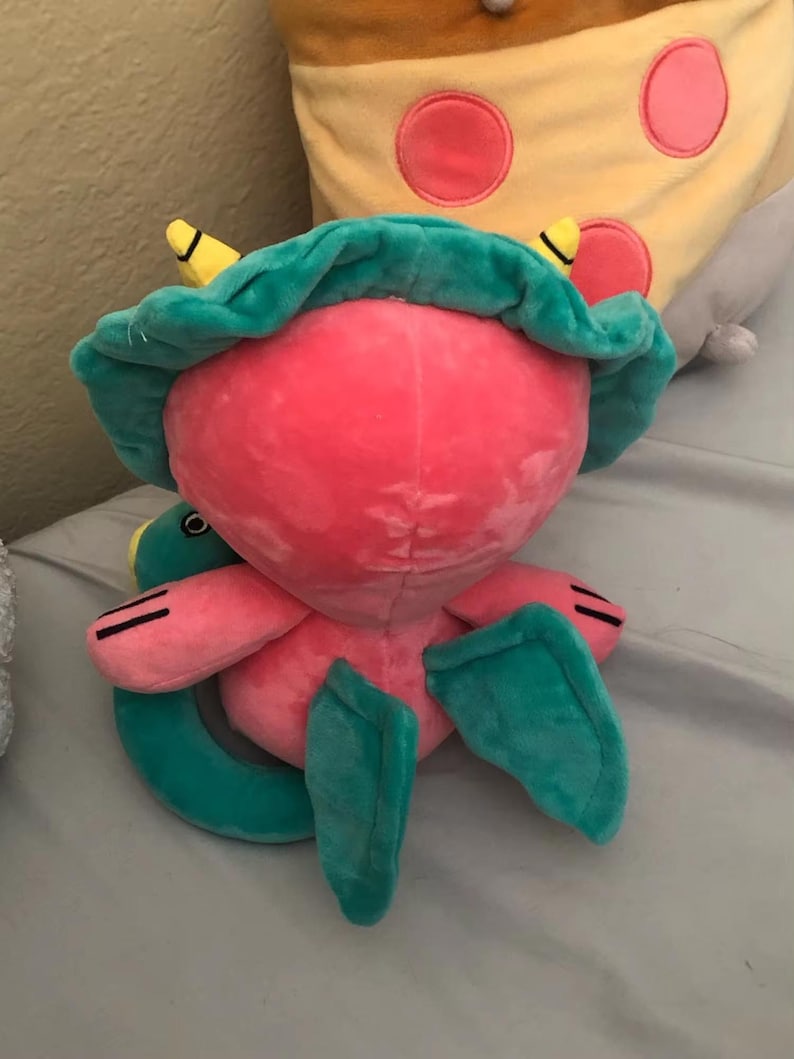 Op de afbeelding: Een roze en groen pluche speelgoed met een groot hoofd en een klein lichaam. Het speelgoed heeft een groene hoed met gele accenten en een lange, gekrulde staart.
