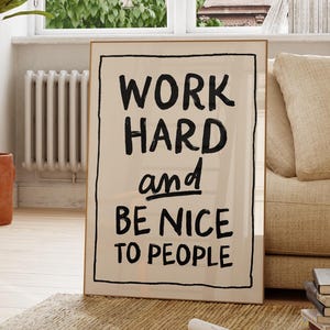 以下が含まれることがあります： 黒い文字で「WORK HARD and BE NICE TO PEOPLE」と書かれた額入りのプリント。 大きな窓、鉢植えの植物、木製の椅子のある、日当たりの良い部屋のベージュのソファにプリントが立てかけられています。