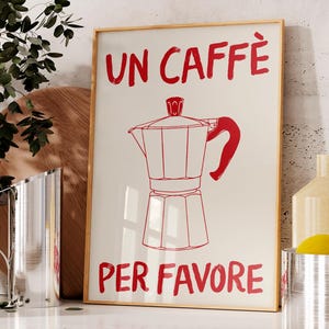 Puede incluir: Un cuadro enmarcado con la frase italiana "UN CAFFÈ PER FAVORE" en rojo, junto con un dibujo lineal rojo de una cafetera italiana. El cuadro está expuesto en una encimera de cocina.