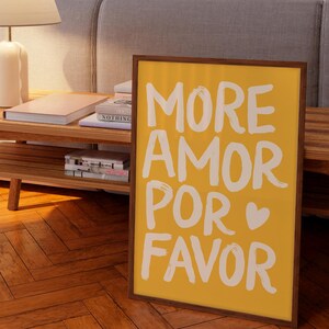 Peut inclure: Int&eacute;rieur de salon avec une table basse en bois, une lampe et des livres. Une affiche encadr&eacute;e avec les mots "MORE AMOR POR FAVOR" en blanc sur fond jaune est pos&eacute;e contre la table. Une autre affiche encadr&eacute;e avec un c&oelig;ur rose est visible.