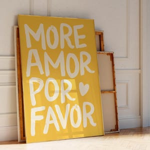 Peut inclure: Une affiche jaune avec l'inscription blanche "MORE AMOR POR FAVOR". L'affiche est appuyée contre un mur blanc, avec un parquet au premier plan. Les mots se traduisent par "Plus d'amour s'il vous plaît".