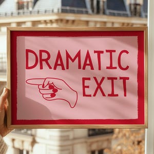 May include: A framed print with the words "DRAMATIC EXIT" in red, and a hand pointing right. The print has a pink background and a red border, set in a light brown wooden frame.
