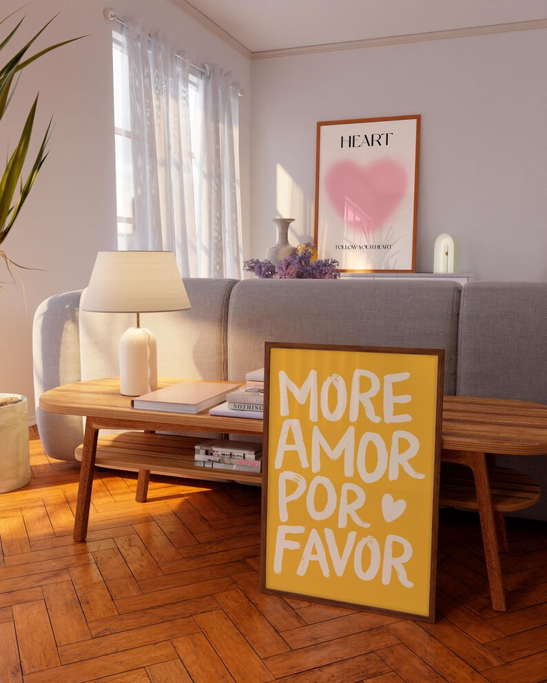 Peut inclure: Int&eacute;rieur de salon avec une table basse en bois, une lampe et des livres. Une affiche encadr&eacute;e avec les mots "MORE AMOR POR FAVOR" en blanc sur fond jaune est pos&eacute;e contre la table. Une autre affiche encadr&eacute;e avec un c&oelig;ur rose est visible.