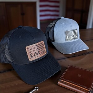 May include: Two baseball caps with mesh backs and leather patches. One is black, the other gray. The patches display an American flag design with the words "FAITH OVER FEAR." A brown leather wallet and car keys are also on the wooden surface.