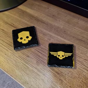 May include: Two square, black coasters with gold skull designs. One coaster features a skull, and the other has a skull with wings. The coasters are made of a dark, textured material and are displayed on a wooden surface.