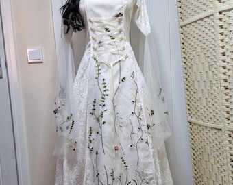May include: A white wedding dress with floral embroidery. The dress features a corset-style bodice with lace-up detailing and long, sheer sleeves. The skirt is floor-length, and the dress is displayed on a mannequin with a black wig.