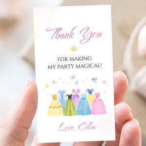 Może przedstawiać: Biała karta z napisem "Thank You" i "For making my party magical!" w różowym skrypcie. Karta zawiera akwarelowe ilustracje sukienek księżniczek w różnych kolorach, z motylami i gwiazdami. Karta jest trzymana przez osobę.