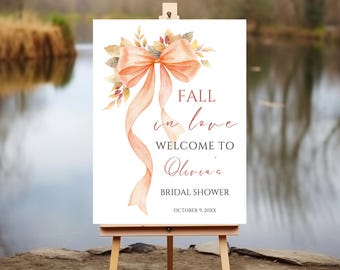 May include: A white sign on a wooden easel with the text "FALL in love WELCOME TO Olivia's BRIDAL SHOWER" in elegant script. The sign is decorated with a watercolor bow and foliage in shades of peach, orange, and gold. The date "OCTOBER 9, 20XX" is at the bottom.