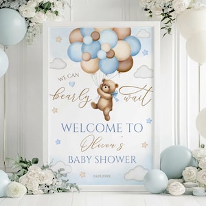 May include: A framed baby shower sign with a teddy bear holding balloons. The sign reads "We can bearly wait" and "Welcome to Oliver's Baby Shower" in blue and gold lettering. The date 04.19.20XX is also included.