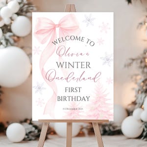 May include: A welcome sign on a wooden easel with a pink bow and the words "Welcome to Olivia's Winter Onerland First Birthday" in elegant script. The sign features snowflakes and a pink Christmas tree.