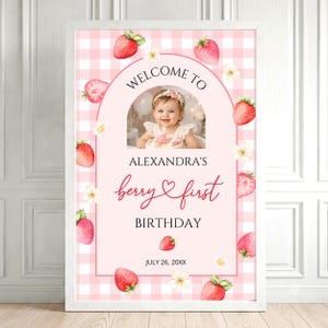 May include: A rectangular birthday sign featuring a pink and white gingham background with watercolor strawberries and white flowers. The center displays a photo of a child, the text "WELCOME TO ALEXANDRA'S berry first BIRTHDAY JULY 26, 20XX."