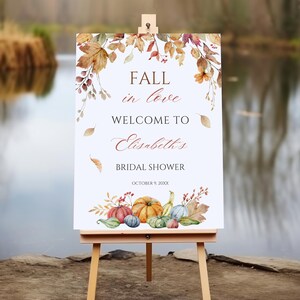 Może przedstawiać: Biały szyld na drewnianym sztaludze z napisem "FALL in love WELCOME TO Elisabeth's BRIDAL SHOWER OCTOBER 9, 20XX." Szyld zdobią akwarelowe dynie, liście i elementy kwiatowe w jesiennych kolorach.