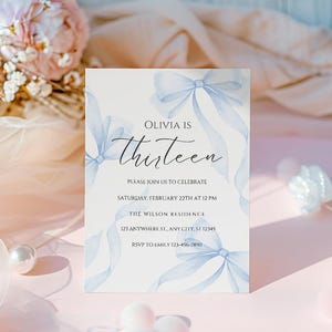 May include: A blue and white invitation card with a watercolor design of bows. The text on the card reads "Olivia is Thirteen" and includes details for a birthday party.