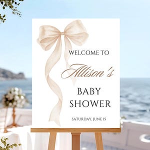 May include: A baby shower welcome sign on a wooden easel. The sign features a watercolour illustration of a beige bow and flowing ribbons. Text reads "Welcome to Allison's Baby Shower" and the date.