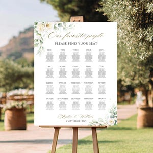 May include: A white wedding seating chart on an easel, with gold script that reads "Our favorite people, Please find your seat." Floral accents and a list of names are displayed. The date "6 September 2025" is at the bottom.