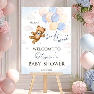 以下が含まれることがあります： 青い風船を持ったテディベアが描かれた、青とピンクのベビーシャワーのウェルカムサイン。サインには「We can barely wait... Welcome to Olivia's Baby Shower June 17, 2023」と書かれています。