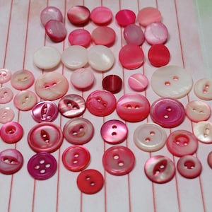 May include: A collection of round buttons in various shades of pink, red, and white. The buttons feature different sizes, textures, and hole configurations, including two-hole and four-hole designs, with some displaying a smooth, pearlescent finish.