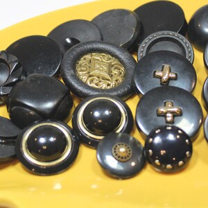 May include: A collection of assorted black buttons in various shapes and sizes, featuring smooth, matte, and glossy finishes. Some buttons include metallic gold accents, intricate embossed patterns, or central studs, while others are plain circular designs.