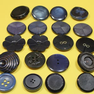 May include: A collection of various dark blue and black buttons in different shapes and designs. Some buttons are round, others are flower-shaped, and some have textured surfaces. The buttons are arranged on a yellow background.