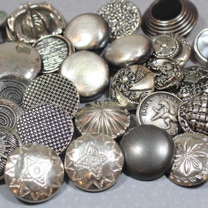 May include: A collection of silver-toned buttons in various sizes and designs. The buttons feature embossed patterns, including floral, geometric, and textured surfaces. Some buttons have a skull, star, or lion design.