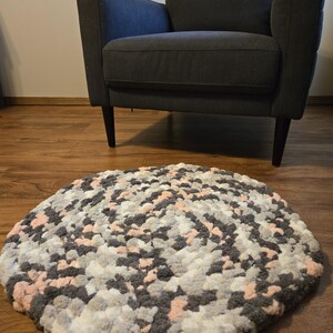 May include: A round, handmade rug with a textured, plush appearance. The rug features a mix of gray, white, and light pink yarn, creating a soft, inviting aesthetic. The rug is placed on a wooden floor, with a gray armchair in the background.