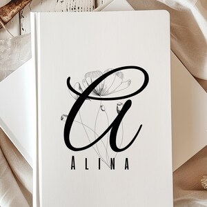 Könnte beinhalten: Ein weißes Notizbuch mit einem schwarzen Blumendesign und dem Namen "Alina" darunter. Das geschlossene Notizbuch liegt auf einer weißen Oberfläche, mit einer Kaffeetasse im Hintergrund.