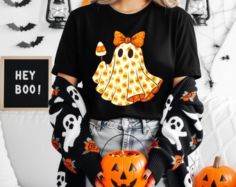 May include: Black Halloween t-shirt with a candy corn ghost design. The ghost has a bow and is holding a candy corn. The person is wearing a black and white ghost-themed cardigan and holding a jack-o'-lantern bucket. The sign says "HEY BOO!"